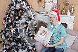Young man with big present sitting on couch near christmas tree and fireplace