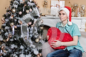 Young man with big red heart sitting on couch near christmas tree and fireplace