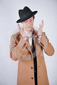 Young man in a black business suit and beige fashion coat with a stylish hat stands in police handcuffs on a white solid Studio ba