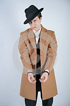 Young man in a black business suit and beige fashion coat with a stylish hat stands in police handcuffs on a white solid Studio ba