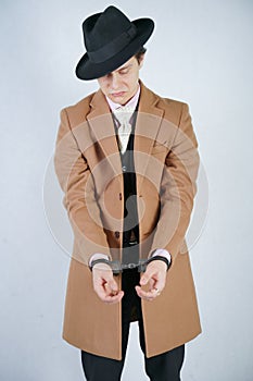 Young man in a black business suit and beige fashion coat with a stylish hat stands in police handcuffs on a white solid Studio ba
