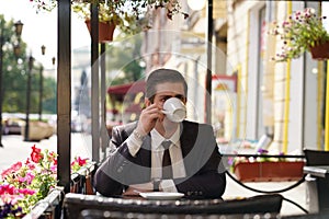 A young man in a black business suit, white shirt and tie sits in a city street cafe at a table and enjoys his cappuccino with foa