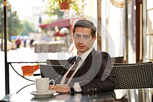 A young man in a black business suit, white shirt and tie is sitting in a city street cafe at a table and enjoying a Cup of coffee