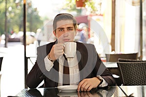 A young man in a black business suit, white shirt and tie sits in a city street cafe at a table and enjoys his cappuccino with foa