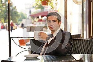 A young man in a black business suit, white shirt and tie sits in a city street cafe at a table and enjoys his cappuccino with foa
