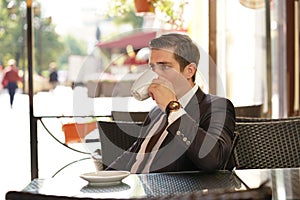 A young man in a black business suit, white shirt and tie sits in a city street cafe at a table and enjoys his cappuccino with foa