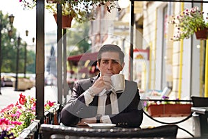 A young man in a black business suit, white shirt and tie sits in a city street cafe at a table and enjoys his cappuccino with foa