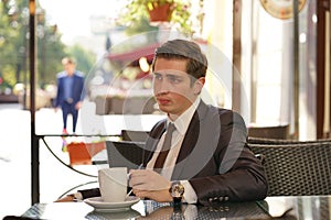 A young man in a black business suit, white shirt and tie is sitting in a city street cafe at a table and enjoying a Cup of coffee