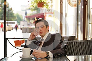 A young man in a black business suit, white shirt and tie is sitting in a city street cafe at a table and enjoying a Cup of coffee