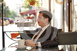 A young man in a black business suit, white shirt and tie is sitting in a city street cafe at a table and enjoying a Cup of coffee