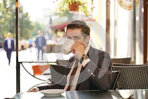 A young man in a black business suit, white shirt and tie is sitting in a city street cafe at a table and enjoying a Cup of coffee