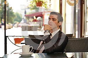 A young man in a black business suit, white shirt and tie is sitting in a city street cafe at a table and enjoying a Cup of coffee