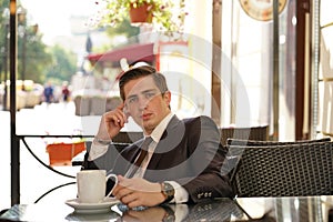 A young man in a black business suit, white shirt and tie is sitting in a city street cafe at a table and enjoying a Cup of coffee