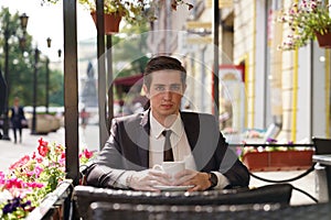 A young man in a black business suit, white shirt and tie is sitting in a city street cafe at a table and enjoying a Cup of coffee