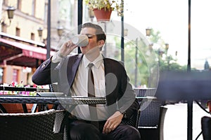 A young man in a black business suit, white shirt and tie is sitting in a city street cafe at a table and enjoying a Cup of coffee