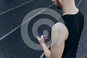 A young man in black clothes is looking at the phone. fitness athlete on the sports field. training with gadgets. warm up body
