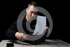 Young man in black hoodie inserts paper into typewriter on black background in low key