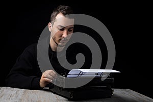 Young man in black hoodie inserts paper into typewriter on black background in low key