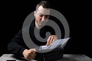 Young man in black hoodie inserts paper into typewriter on black background in low key