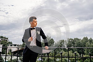 a young man in a black jacket with a bud of flowers stands on the balcony. groom