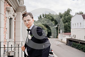 a young man in a black jacket with a bud of flowers stands on the balcony. groom