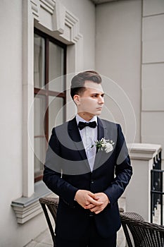 young man in a black jacket with a bud of flowers stands on street near building