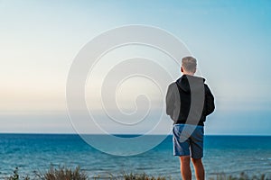 A young man in a black jacket stands with his back to the camera and looks at the blue sea against the background of a calm sunset