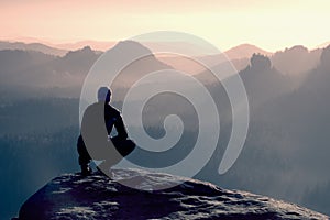 Young man in black sportswear is sitting on cliff's edge and looking to misty valley bellow