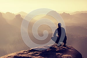 Young man in black sportswear is sitting on cliff's edge and looking to misty valley bellow