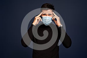 Young man in black sweater, in sterile face mask feeling sick or dizzy, touching head, suffering headache or fever from