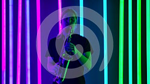 A young man in a black T shirt plays the saxophone in a dark studio against a background of bright neon lights. Slow