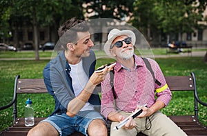 Young man and blind senior sitting on bench in park in city, using smartphone.