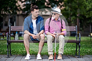 Young man and blind senior with white cane sitting on bench in park in city.