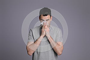 Young man blowing nose in tissue on grey background. Cold symptoms