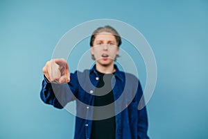 Young man on a blue background points at you. Surprised guy in a blue shirt stands against the wall background and points his