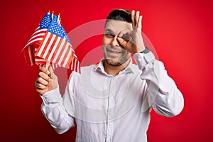 Young man with blue eyes holding flag of united states of america over red isolated background with happy face smiling doing ok