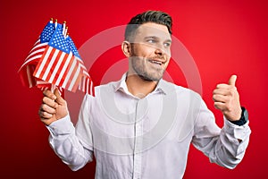 Young man with blue eyes holding flag of united states of america over red isolated background pointing and showing with thumb up