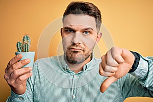 Young man with blue eyes holding fresh cactus plant pot over yellow background with angry face, negative sign showing dislike with