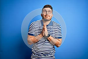 Young man with blue eyes wearing glasses and casual striped t-shirt over blue background begging and praying with hands together