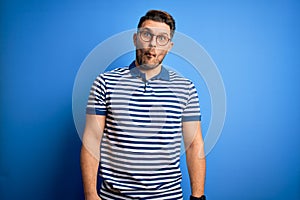 Young man with blue eyes wearing glasses and casual striped t-shirt over blue background making fish face with lips, crazy and