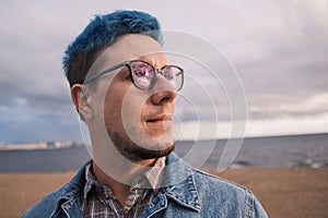 A young man with blue hair and glasses looks at the sunset.
