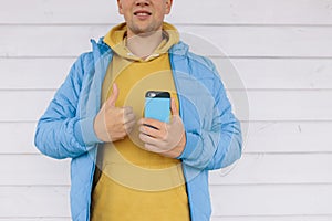 Young man in a blue jacket holding a mobile phone in one hand, and the other showing a thumbs up