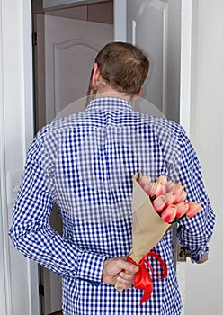 A young man in a blue plaid shirt and jeans, holding a bouquet of tulips behind his back, and peeks in the open door