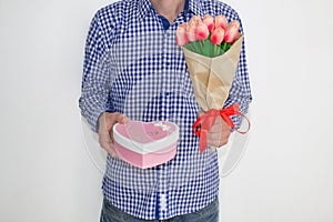 A young man in a blue plaid shirt and jeans, holding a bouquet of tulips and a gift box in the shape of a heart, on a white backgr