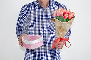 A young man in a blue plaid shirt and jeans, holding a bouquet of tulips and a gift box in the shape of a heart, on a white backgr