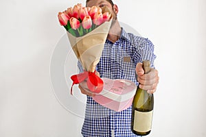 A young man in a blue plaid shirt stretches a bouquet of tulips, a heart-shaped gift box and a bottle of wine