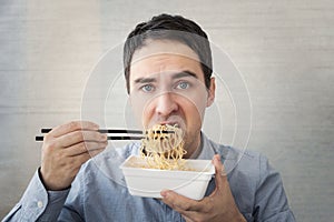 young man in a blue shirt is eating noodles from a box with a dissatisfied face. Lunch at the office. tasteless junk