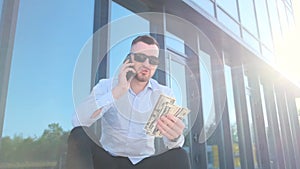 A young man in a blue shirt sits on the steps in front of the office with dollars in his hands