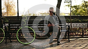 Young man in blue shirt sittting on the bench and holding his mobile phone in hands, texting. Rider taking a rest after