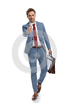 Young man in blue suit holding suitcase and making thumbs up gesture